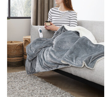 Een vrouw zit onder het SleepMed Elektrische Knuffeldeken in de lichtgrijs. 