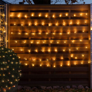 Een hek en een bosjes zijn versierd met de netverlichting. De lampjes geven warm wit licht. 