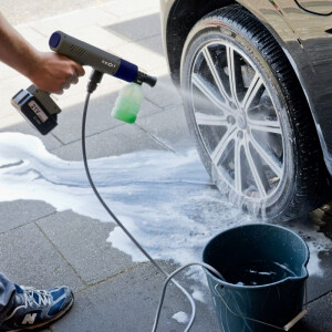 Nexxt Draadloze Hogedrukreiniger Deluxe in gebruik bij het reinigen van een autoband met schuimlans en emmer water op de oprit.